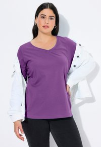 Fialové tričko s výstřihem do V vyrobené z měkké tkaniny, s krátkými rukávy a volným střihem, doplněné bílou bundou přehozenou přes ramena.