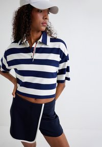 Polo à manches courtes rayé bleu marine et blanc avec un col et des boutons ; associé à un short sombre avec une bordure blanche.