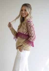 Beżowy sweter dzianinowy z różowym geometrycznym wzorem i frędzlami na rękawach. Modelka ma na sobie białe spodnie, eksponując wzór swetra.