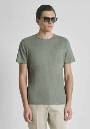 Muškarac koji nosi tamne sunčane naočale, kratku majicu maslinasto zelene boje i bež hlače, stoji ispred jednobojne svijetle pozadine.