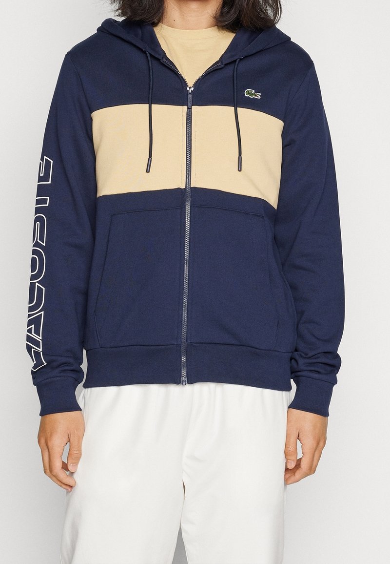 Felpa con zip navy e pannello orizzontale beige. Dotata di cappuccio regolabile, tasche frontali e logo sulla manica sinistra. Tessuto morbido.