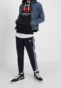 Giacca di jeans, felpa nera con cappuccio e grafica, t-shirt bianca, pantaloni da tuta blu navy con striscia laterale e sneakers nere alte.