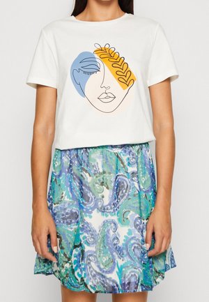 Femme portant un t-shirt blanc avec un dessin abstrait de visage en lignes bleues et orange, associé à une jupe à motif cachemire bleu et vert.