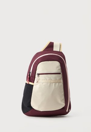 Sac à dos bandoulière compact en bordeaux, beige et noir avec poches avant zippées et une sangle rembourrée sur fond uni.