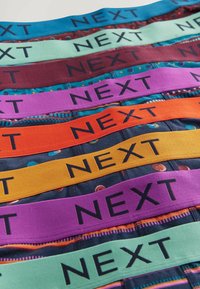 Cinturilla elástica en múltiples colores: azul, verde, púrpura, rojo, naranja y amarillo, todos con "NEXT" impreso en negro. Tejido texturizado.
