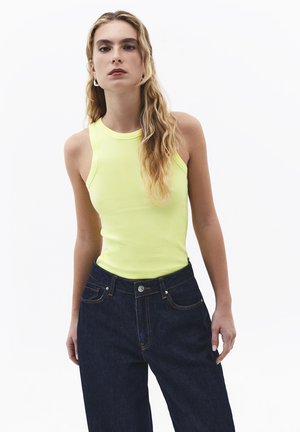 Jeune femme aux longs cheveux blonds ondulés, portant un haut jaune sans manches et un jean taille haute bleu foncé sur fond blanc.