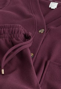 Burgundy ribbed cardigan with rounded neckline, two buttons, and a front pocket. Soft texture with an elastic waistband detail.