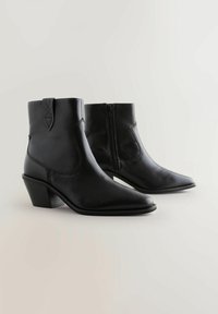 Botas de cuero negras hasta el tobillo con punta afilada, cremallera lateral y tacón curvado. Presentan detalles de costura y un acabado suave.