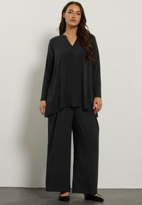 Tunic nera a maniche lunghe con scollatura a V e orlo a strati; abbinata a pantaloni neri a gamba larga e scarpe nere con tacco basso. Texture liscia.