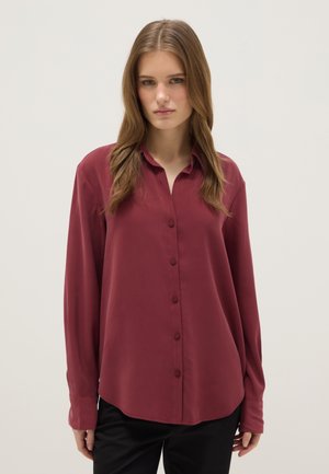 Junge Frau mit langen Haaren trägt ein locker sitzendes dunkelrotes Hemd mit Knöpfen und schwarze Hose, steht vor einem schlichten hellen Hintergrund.