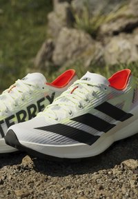 Λευκά και μαύρα παπούτσια τρεξίματος Adidas Terrex με κορδόνια νέον κίτρινου χρώματος και πορτοκαλί εσωτερική επένδυση σε βραχώδες έδαφος σε εξωτερικό χώρο.