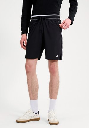 Mężczyzna w czarnych sportowych szortach ze sznurkiem i kieszenią na zamek, w białych skarpetkach i białych trampkach, stojący na jednolitym tle.