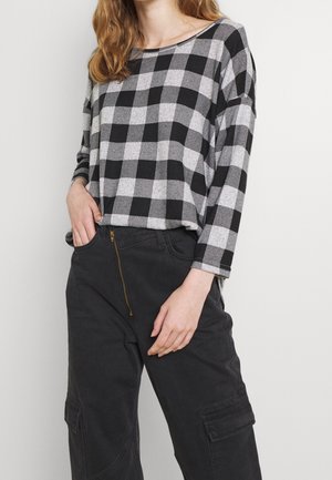 Camicia a maniche lunghe a quadri neri e grigi con scollatura ampia, abbinata a pantaloni cargo scuri con cerniera dorata e dettagli delle tasche.
