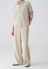 Chemise beige rayée avec une poche, assortie à un pantalon large dans le même tissu. Présente une coupe décontractée et des manches retroussées.