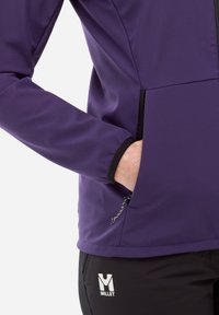 Main dans la poche zippée latérale d'une veste violette portée avec un pantalon noir arborant un logo Millet blanc sur la cuisse.