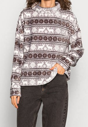 Personne portant un pull en polaire gris et blanc à motifs de rennes et de flocons de neige, associé à un jean gris foncé.