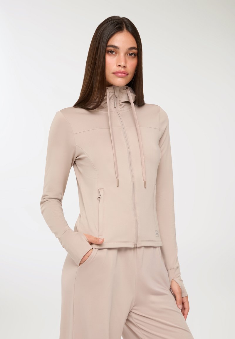 Giacca beige chiaro con zip realizzata in tessuto morbido e stretch. Presenta un colletto alto, cordoncino con fermagli in metallo e tasche laterali.