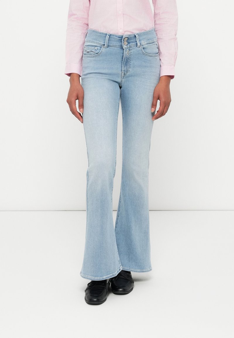 Jean flare bleu clair avec une taille haute, dotés de poches avant et d'une fermeture éclair classique. Portés avec des chaussures noires.