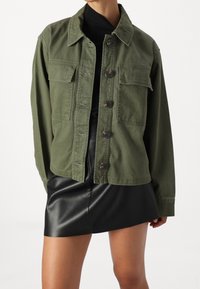 Olivově zelená džínová bunda s límcem, knoflíkovým zapínáním a dvěma kapsami na hrudi. Má volný střih a texturovanou látku.