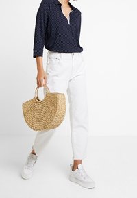 Navy-blaues Polka-Dot-Shirt mit Kragen, weiße hochtaillierte Jeans und eine gewebte Strohtasche. Weiße Sneakers mit dicker Sohle.