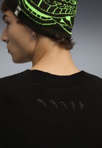 Jersey negro con cuello acanalado, que presenta gráficos sutiles bordados en negro en la parte posterior. Tejido de punto con textura visible.