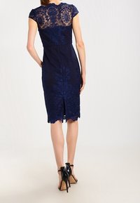 Navy mekko, jossa on pitsinen yläosa hihatettömillä hihoilla, monimutkaisia kukkakuosisia yksityiskohtia ja sakarat helmaa. Takareunassa halkio liikkuvuuden helpottamiseksi.