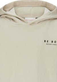 Felpa con cappuccio beige con il testo nero "BE BO" e un testo più piccolo "RETOUR SINCE" nella parte superiore sinistra del petto.