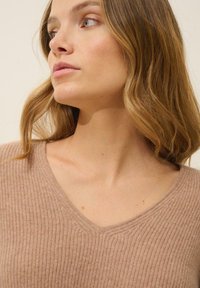 Beżowy, prążkowany sweter z dekoltem w serek. Miękka tekstura, dopasowany krój. Zbliżenie na dekolt i część obojczyka widoczna.