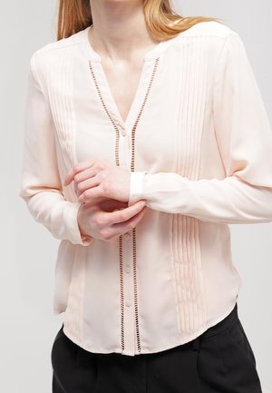 Frau trägt eine langärmelige, hellrosa Bluse mit vertikalen Falten und Cut-out-Details, hält ein Handgelenk mit der anderen Hand.