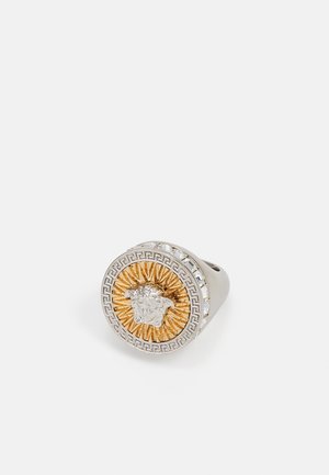 Bague en argent et or, mettant en avant une tête de lion au centre, entourée d'un motif en rayons de soleil et d'un motif de clé grecque, avec des accents en cristal.