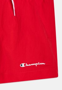 Shorts de bain rouges en tissu léger, dotés d'une taille à cordon de serrage et d'un logo "Champion" blanc en bas à gauche.