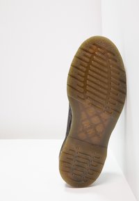 Semelle d'une chaussure foncée avec un motif de bande de roulement en caoutchouc translucide marron et un texte en relief, positionnée verticalement contre un mur d'angle blanc.