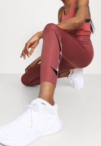 Donna in ginocchio con leggings Nike color bordeaux e canotta, indossando scarpe e calze bianche Nike su uno sfondo semplice.
