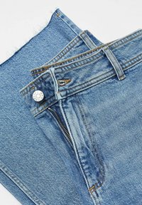 Jean en denim bleu plié, montrant une fermeture éclair avant partiellement ouverte, un bouton en métal et des passants de ceinture avec des détails de couture visibles.