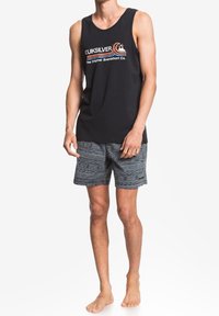 Svart linne med en vit och röd "Quiksilver"-logga, tillsammans med mönstrade grå badshorts med geometriska mönster. Barfota.