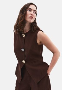 Bruine linnen gilet met een ronde halslijn en zonder mouwen, voorzien van decoratieve knopen in goud, zilver en crème. Inclusief zijzakken.