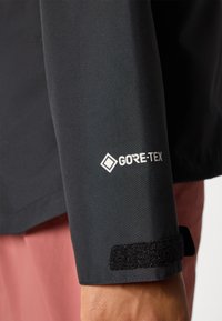 Črna jakna z rokavom, ki ima belo logo "GORE-TEX" in Velcro trak, z zavihkom roza tkanine pod njim.