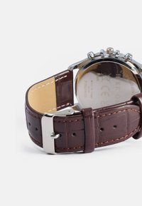 Brūns ādas pulksteņa siksniņa ar krokodila rakstu, sudraba nerūsējošā tērauda sprādze un pulēta nerūsējošā tērauda aizmugure.