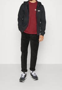 Marinblå zip-up hoodie över en vinröd t-shirt, svarta byxor och mörka sneakers med vita snören. Har frampocketar på hoodien.