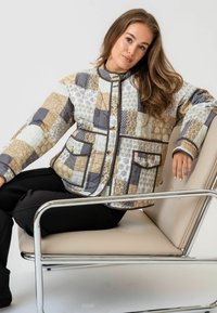 Jachetă matlasată cu un design patchwork în nuanțe de bej, gri și alb, cu butoane rotunde și buzunare laterale. Material neted și ușor.