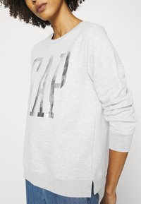 Sudadera gris con un logo metálico de "GAP", cuello redondo, mangas largas y aberturas laterales en el dobladillo para mejorar el ajuste y la comodidad.