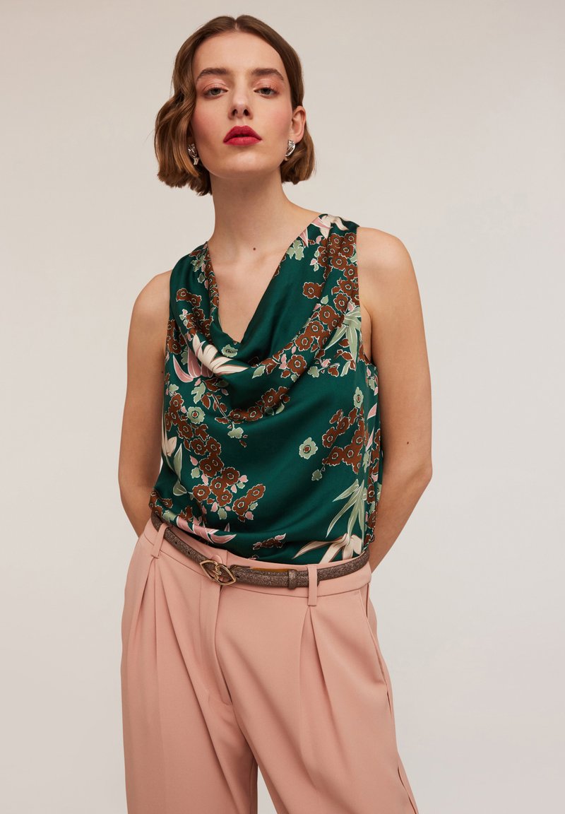 Beżowy top bez rękawów w zielonym kolorze z kwiatowym wzorem; z dekoltem typu cowl. Połączony z jasnoróżowymi spodniami, z fałdami i dekoracyjnym paskiem.