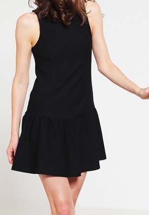 Kvinde iført en ærmeløs sort kjole med tætsiddende overdel og udvidet skørt, stående mod en ensfarvet hvid baggrund.