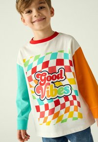 T-shirt a maniche lunghe di colore bianco, con un motivo a quadretti colorato in blu, rosso e verde. Testo "Good Vibes" in grassetto, con un font giocoso.