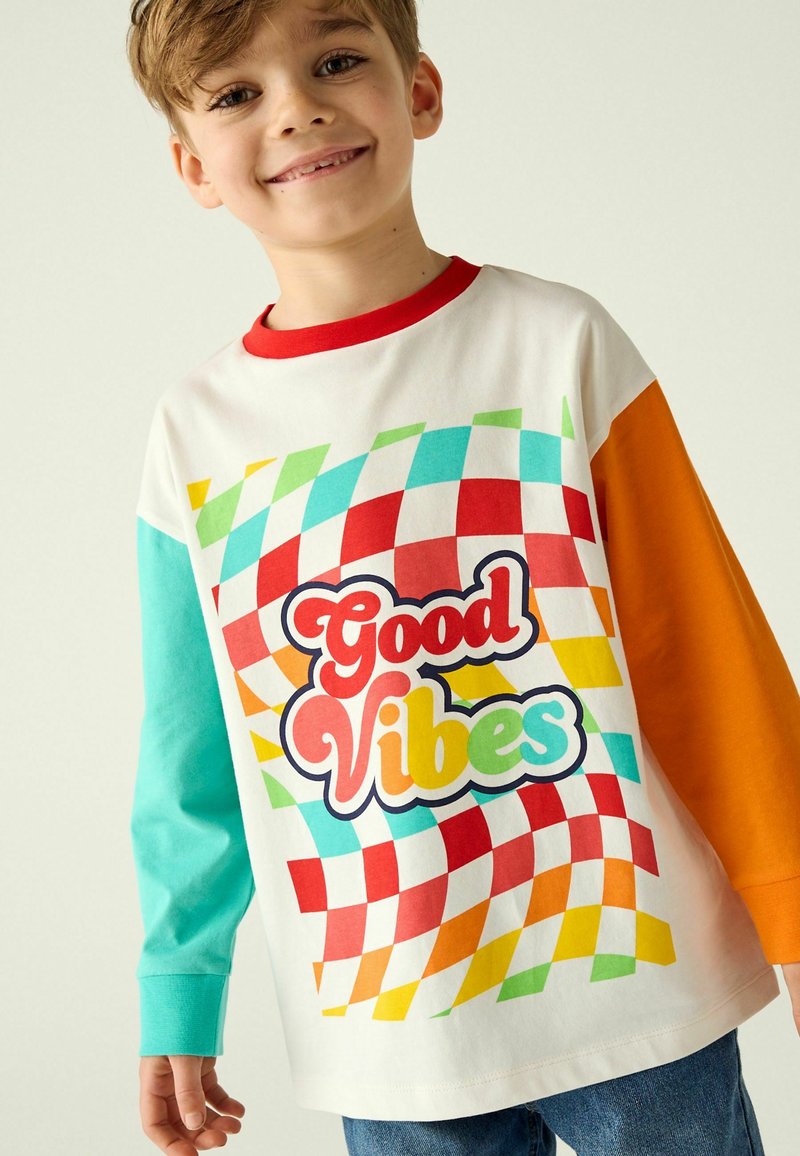 T-shirt a maniche lunghe di colore bianco, con un motivo a quadretti colorato in blu, rosso e verde. Testo "Good Vibes" in grassetto, con un font giocoso.
