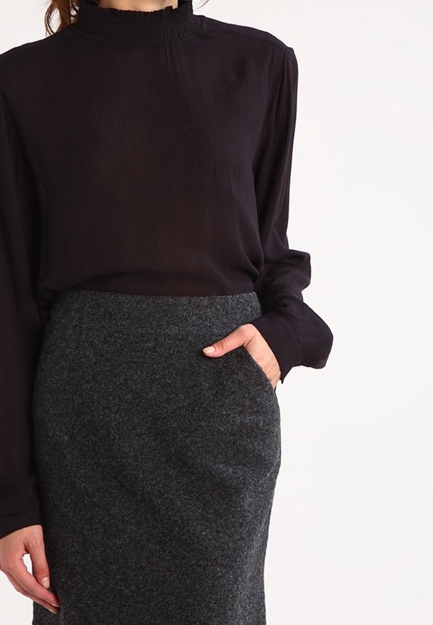 Schwarze Bluse mit langen Ärmeln und hohem Rüschensaum, kombiniert mit einem knielangen grauen Wollrock mit seitlichen Taschen.