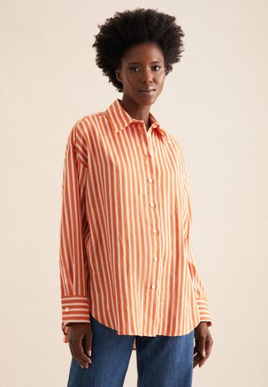 Chemise à manches longues rayée orange et blanche avec un bouton devant, un col évasé et une coupe légèrement ample, associée à un jean en denim.