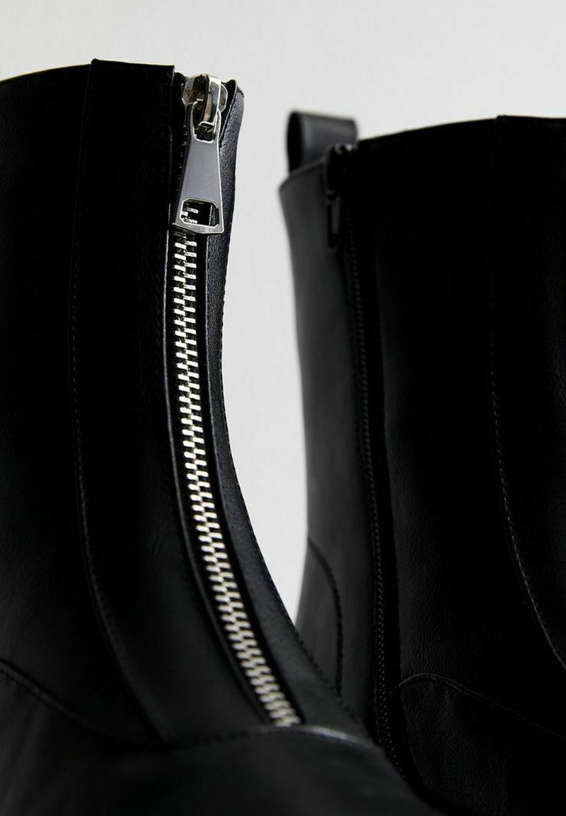 Bottes à chevilles en cuir noir avec des fermetures éclair argentées, présentant une texture lisse et une forme structurée, conçues avec une fermeture éclair sur le côté pour un accès facile.