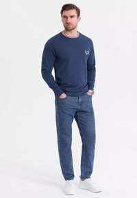 Navy long-sleeve sweater with red and white striped collar, logo accent. Paired with light blue jeans and white sneakers. Casual fit.