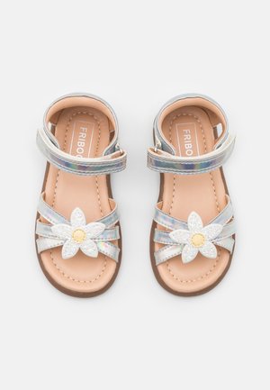 Sandalias iridiscentes con un adorno floral, que presentan una flor blanca con centro amarillo. Las correas son plateadas, con una plantilla marrón claro y suela marrón.